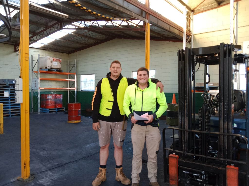 Jake and Bryce from Nexus standing in a warehouse near a forklift - Bryce proudly holds his forklift license.