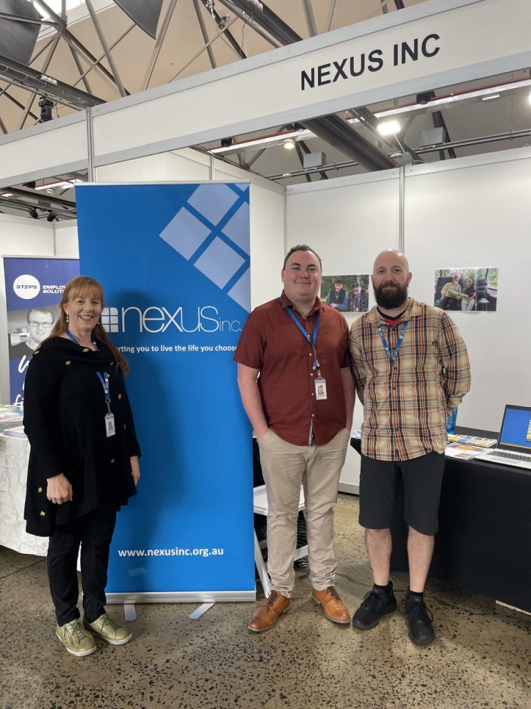 Three Nexus staff standing next to the Nexus banner and booth at the 2022 Speak Out Disability Expo