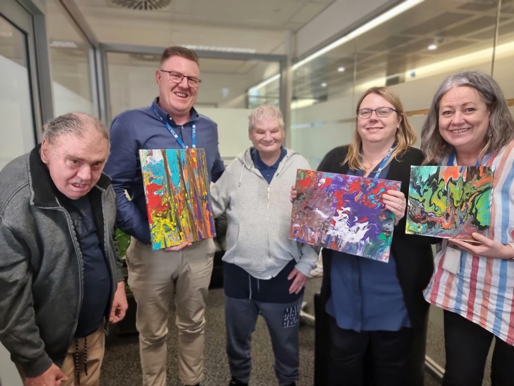 A group of smiling Nexus staff pose with two clients and the beautiful artworks they have created.