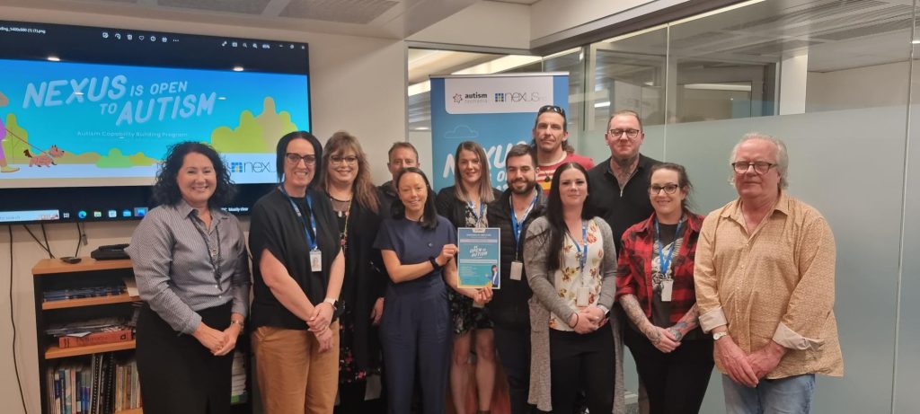 A group of 12 people stand in front of a banner that says Nexus is Open to Autism at the graduation of staff from the Autism Capability Building Program