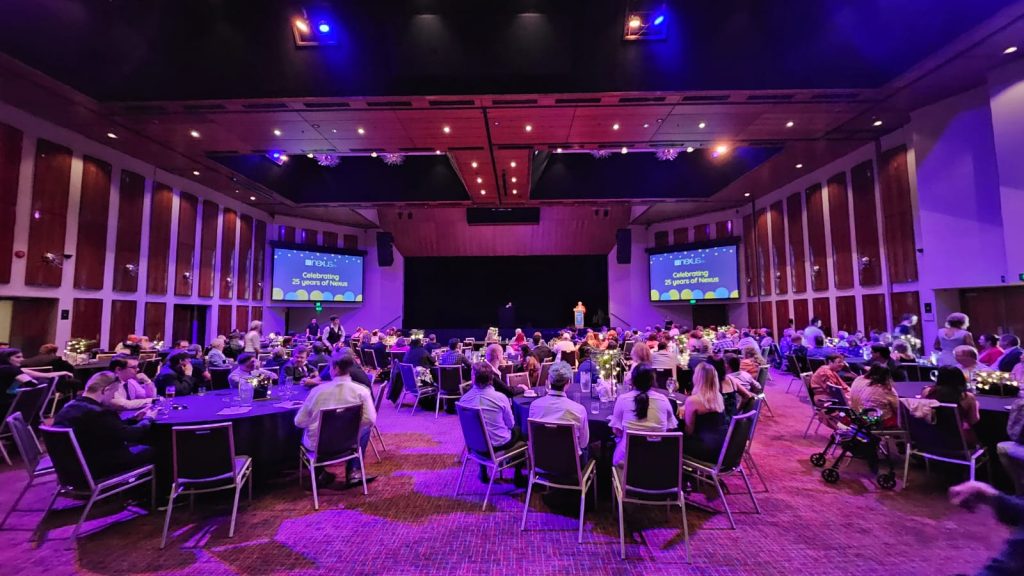 Formal dinner set up, a selection of round tables with guests looking toward the stage and two big screens welcoming everyone to the Nexus 25th Birthday event